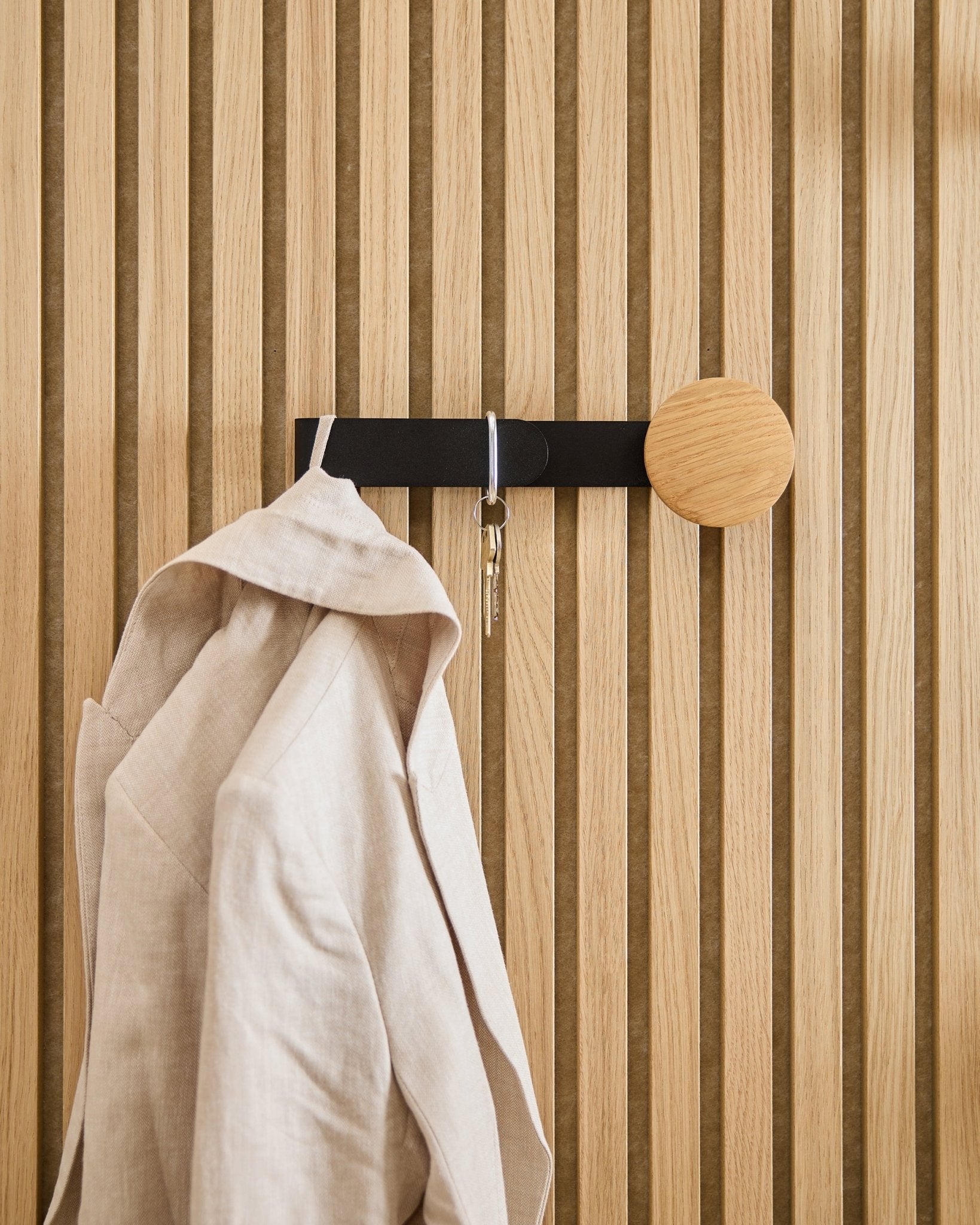 Un blazer beige e un mazzo di chiavi sono appesi all’appendiabiti WoodUpp Linn Horizontal in natural oak, installato su un pannello acustico in legno natural oak Akupanel.